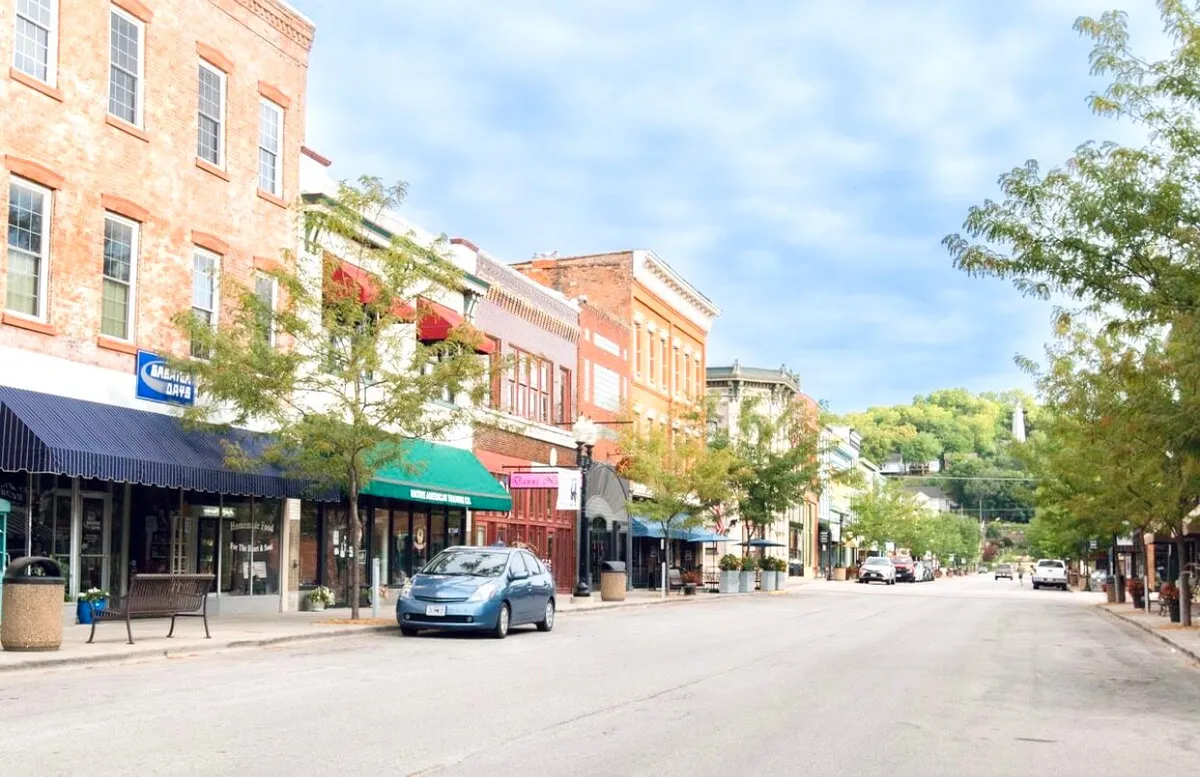 A lively downtown street scene featuring historic buildings, colorful storefronts, leafy trees lining the sidewalk, and parked cars. The area is bustling, evoking a sense of local activity and community, perfect for festival settings.