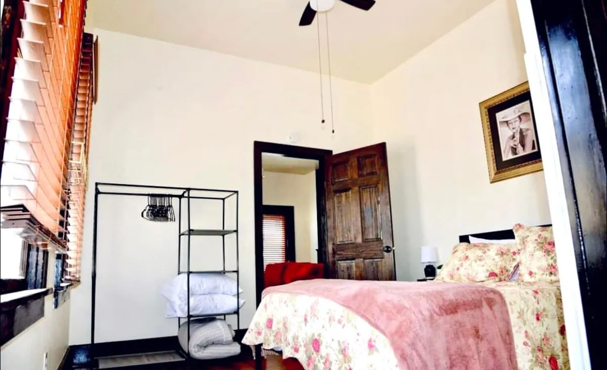 A welcoming bedroom with a bed dressed in floral bedding, an open door leading to an adjoining space, and a shelf stacked with neatly folded towels. The room is bathed in natural light, evoking a cozy, homey feel ideal for guests.