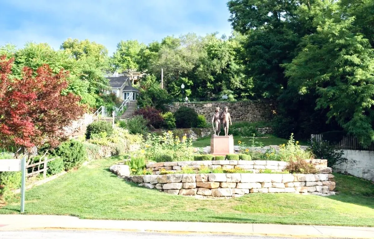 A serene garden area with a sculpture of two figures, surrounded by vibrant flower beds, lush greenery, and a stone retaining wall. The landscaped space invites guests to stroll, relax, or find artistic inspiration outdoors.