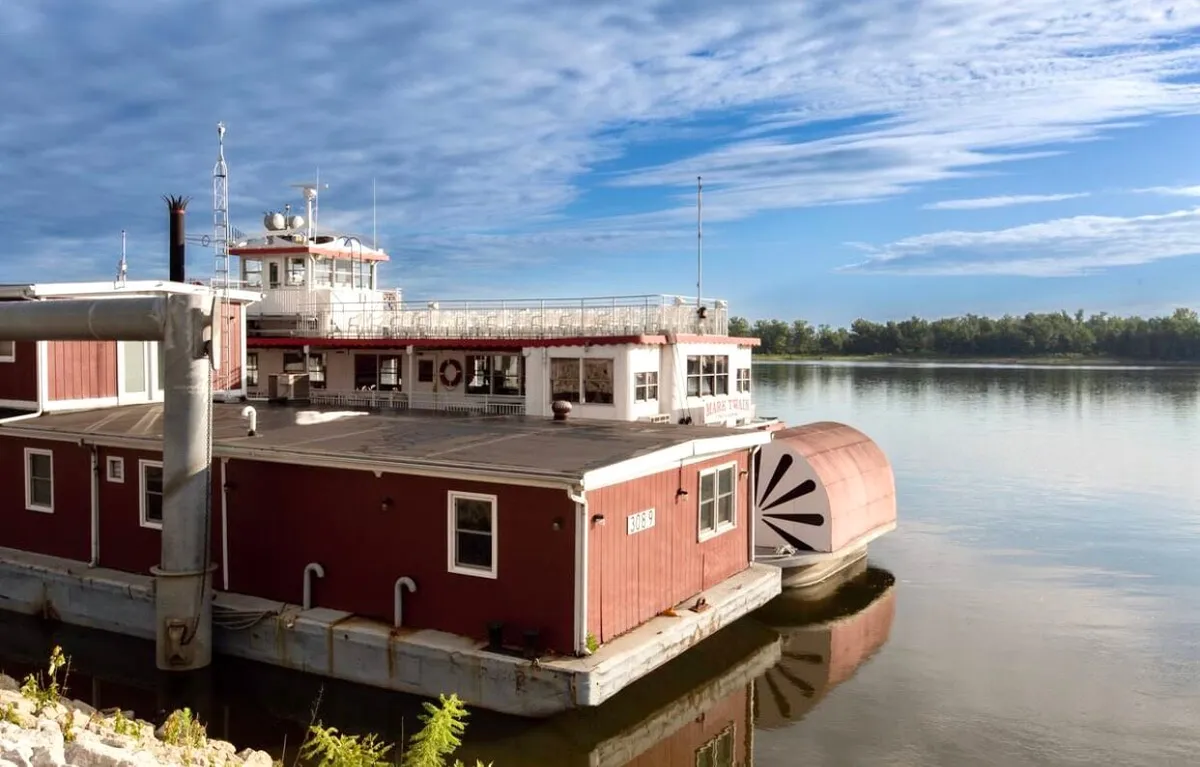 A welcoming exterior view of the houseboat structure overlooking the river, with a spacious deck area perfect for relaxing or entertaining. The tranquil water, sky reflections, and inviting deck suggest opportunities for enjoying the outdoor atmosphere and scenic surroundings.