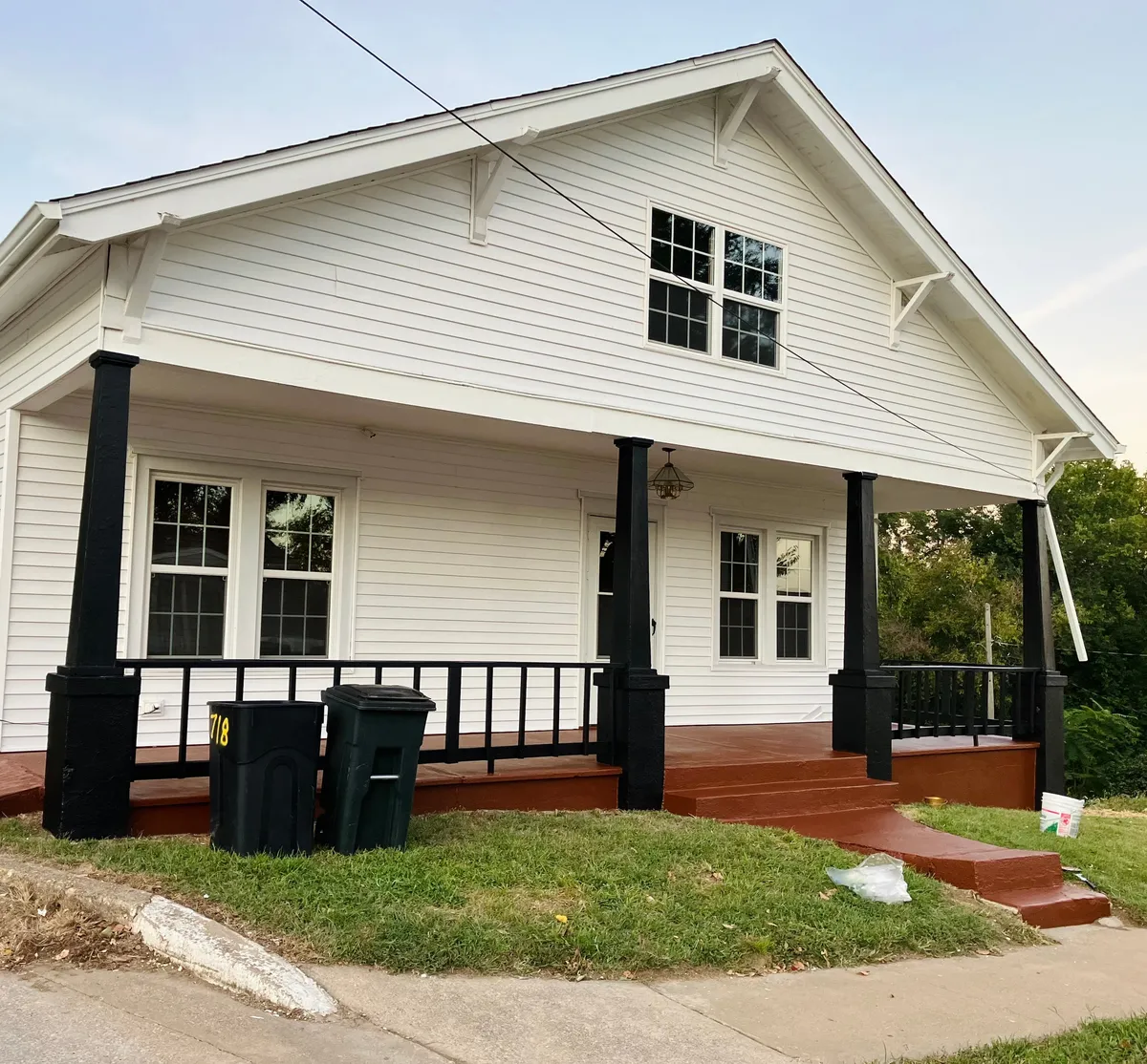 The inviting exterior of Parlor on Paris, with a welcoming front porch framed by bold black pillars and white siding, overlooks green grass. The porch offers a comfortable space for relaxing, socializing, or enjoying the sunset after a day out.