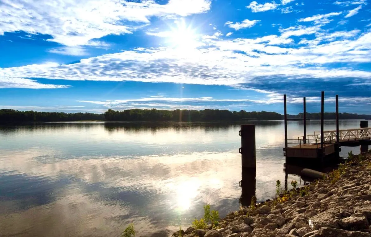 A tranquil riverside view featuring sunlight dancing on the water, a dock extending into the river, and a natural riverbank. The scene encourages exploration, relaxation, or enjoying peaceful moments by the water.