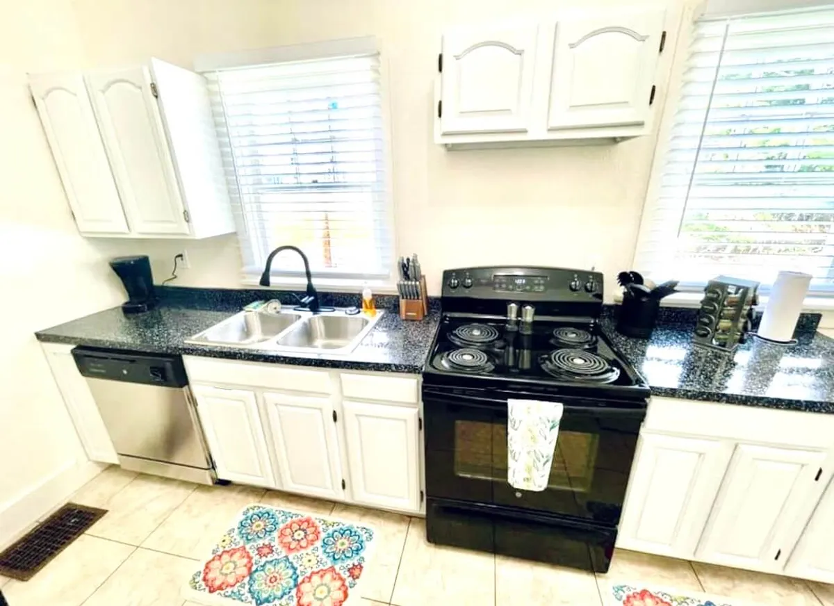 A warm kitchen scene featuring stainless steel appliances, a granite countertop, a colorful floor mat, and a window with blinds. The kitchen is lively and equipped for baking, cooking, or enjoying a morning coffee.