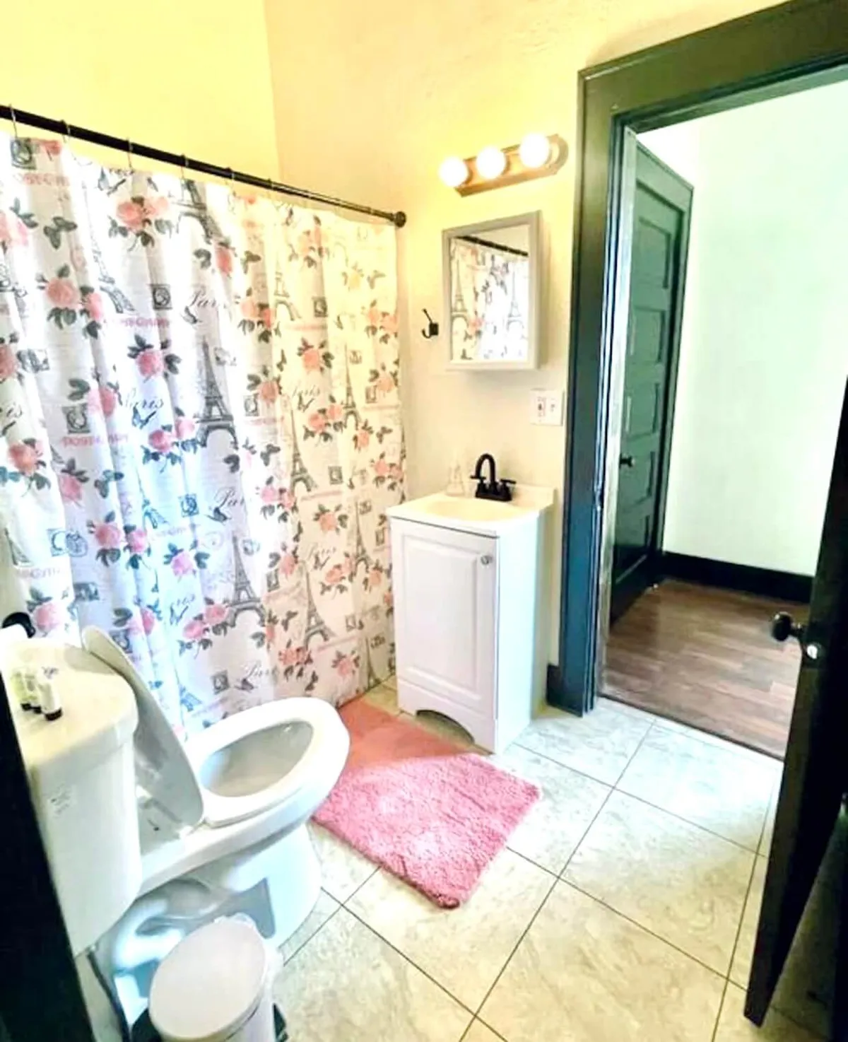 A cheerful bathroom featuring a floral shower curtain, white vanity, pink bath mat, and large mirror. Morning sunlight streams in, casting a bright, fresh glow that makes the space inviting and pristine.