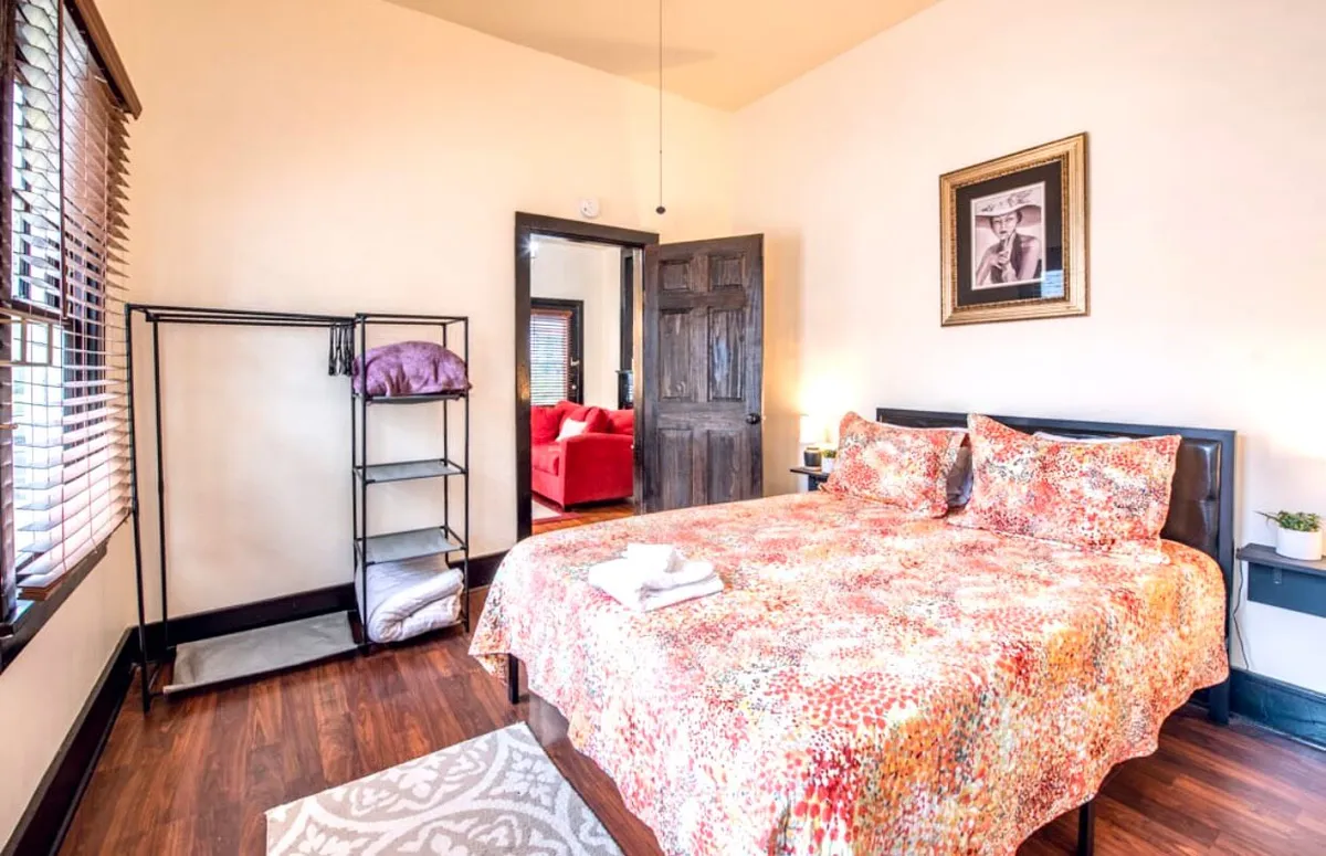 A cozy bedroom features a bed with colorful bedding, decorative wall art above the headboard, and a clothes rack in the corner. Soft natural light filters through unseen windows, creating a warm and restful atmosphere perfect for relaxation or reading.