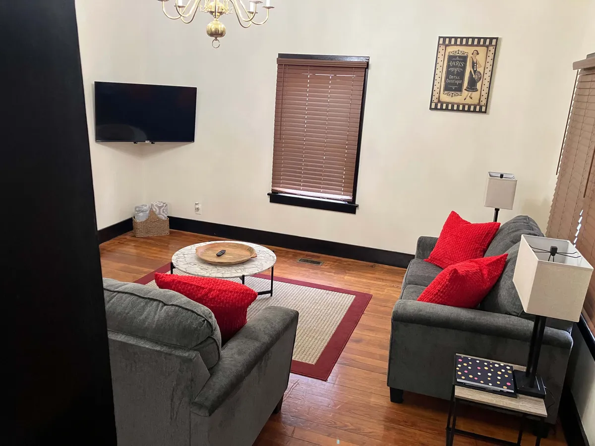 A wide view of the main living area with gray sofas, red accent pillows, a wooden floor, and a coffee table topped with a decorative tray. The space feels open and accommodating, ideal for conversations, watching movies, or sharing laughs with loved ones.